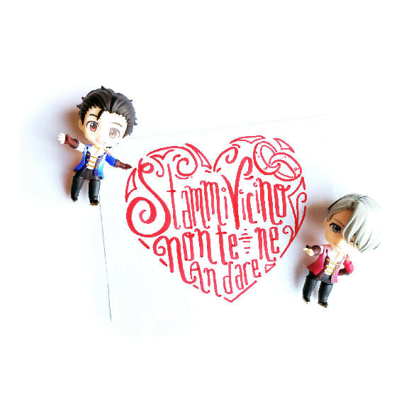 Nendoroids Yuri and Victor next to a red heart. The red heart has the letters "Stammi vicino, non te ne andare" written in it, with a motif of matching rings.